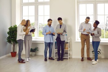 Ofiste çalışan bir grup insan. Karışık ırk melezi erkek ve kadın çalışanlar ve iş adamları iş yerinde bir araya gelip projeleri tartışıyor ve dizüstü bilgisayar ve tablet kullanıyorlar.