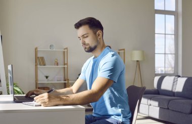 Yetişkin bir adam evden ders çalışıyor ve internetten kurs kullanıyor. Mavi tişörtlü genç bir erkek öğrenci çalışma masasında dizüstü bilgisayar kullanıyor ve not defterine not alıyor. Çevrimiçi eğitim kavramı