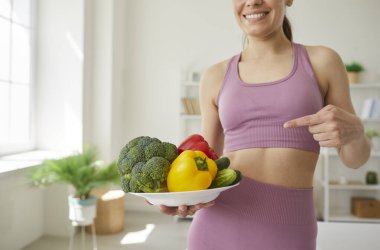 Genç kadın spor yaparak ve taze sağlıklı yiyecekler yiyerek formda kalıyor. Oturma odasında elinde çiğ sebze tabağı olan mutlu, ince bir kız duruyor. Diyet kavramı 