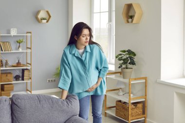 Hamile bir kadın karnına dokunarak doğum sancısı çekiyor. Bekleyen bir kadın, evde tek başına karın ağrısından muzdarip. Hamilelik ve doğum kavramı.