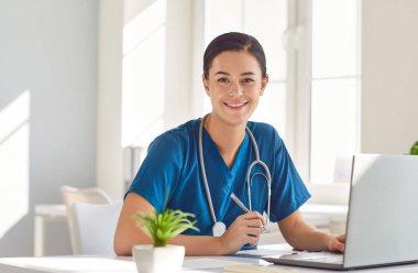 Mavi üniformalı neşeli, dost canlısı, genç bayan doktor portresi bilgisayarda oturuyor ve çalışıyor. Steteskopta gülümseyen kadın doktor kameraya bakıyor, dizüstü bilgisayar kullanıyor ve yazıyor..