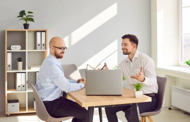 Two smiling businessmen chatting while sitting at laptop in office. Excited caucasian male partners sharing ideas, discussing new project, brainstorming, having informal conversation