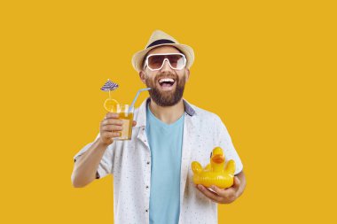 Portrait of a funny bearded man in beach hat and sunglasses holding orange juice cocktail and inflatable duck isolated on yellow studio background. Summer holiday trip and vacation concept