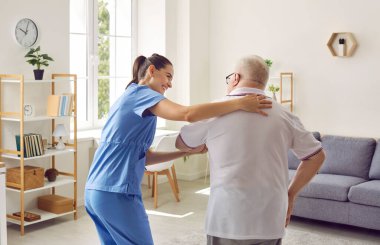 Caring caregiver woman assisting her disabled elderly patient. View from behind of nurse in unifrom helping senior man to walk. Professional medical help and support of elderly people