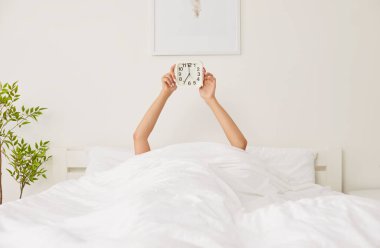 Faceless female person waking up early in the morning lying in her bed under cozy white blanket in modern bedroom holding alarm clock in hands. Good morning, awake and new day concept.