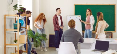 Team of college students presents their project together in classroom near whiteboard. Male teacher sits at desk and listens to his students presenting their work. College teamwork concept.