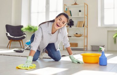 Daily housekeeping routine. Happy young woman wiping dust with spray and rag while cleaning floor at home. Portrait of smiling young Caucasian woman who is cleaning house in her spare time.