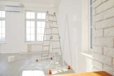 Modern, newly renovated, brightly lit studio apartment. Empty interior with step ladder, freshly painted white walls, bucket, new windows, and protective plastic cover on floor. House renewal concept
