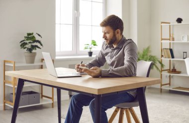 Focused businessman using smartphone and laptop computer at home office. Confident young man in casual wear holding mobile phone and looking at computer screen while sitting at his working place