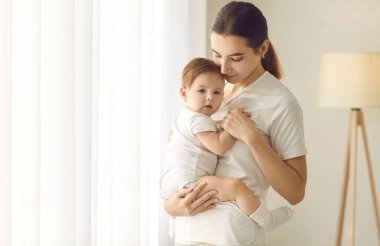 Happy mother together with her child at home. Beautiful young mom standing in a cozy room and holding her cute, sweet baby boy or girl in her arms. Family, love, care, motherhood, and nurture concepts