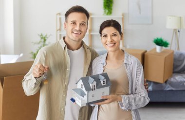 Happy couple of new homeowners holding key and house model. Proud married man and woman smiling and standing in living room, cheerful people celebrating relocation and moving day near cardboard boxes