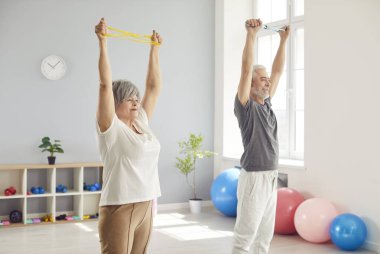 Cheerful senior couple doing sport stretching exercises during arm rehabilitation in clinic. Joyful smiling elderly man and woman training together in gym, working on their mobility and wellbeing.