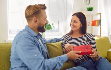 Happy husband expresses love by presenting a gift box, surprise to wife on the home sofa. The image captures the essence of celebration and couple love, symbolizing the joyous occasion.
