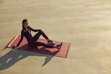 Genç bir kadın şehirde spor yaptıktan sonra dinleniyor. Spor giyimli güzel bir kız gri beton şehir spor sahasında yoga minderinde oturuyor. Spor konsepti. Boşluk arkaplanı kopyala