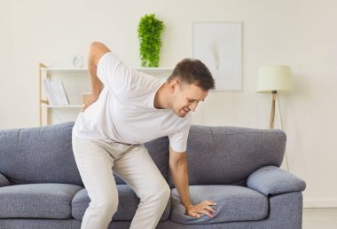 Sick man with lower back pain standing up from sofa in living room at home, holding lumbar with hand. Sad young person suffering from sciatica, pinched nerve or inflammation of intervertebral disc