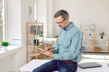 Ciddi sakallı, gömlekli, iş masasında dizüstü bilgisayarlı, cep telefonlu, iş uygulamalı, kısa mesajlı, internetten bilgi arayan olgun bir iş adamı.