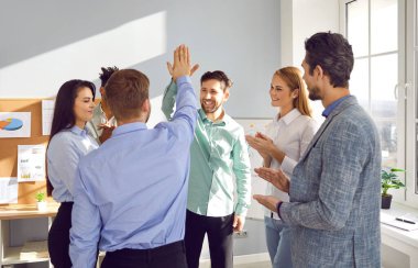 Neşeli iş ekibi başarıyı kutluyor. Mutlu, gülümseyen genç adamlar, meslektaşları alkışlarken birbirlerine beşlik çakıyorlar. Takım çalışması, işbirliği kavramı