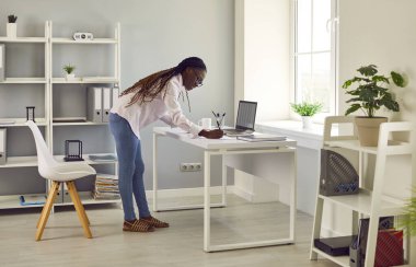 Business woman, corporate employee or financial accountant in modern office workplace. Young African American woman in shirt and jeans standing by work desk with laptop and taking notes on paper