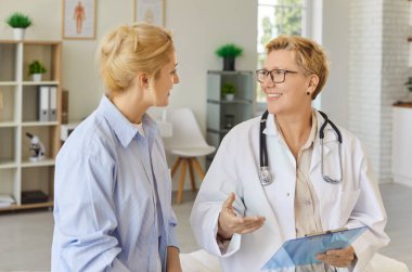 Doctor explaining treatment plan to young smiling patient, medical advice and friendly conversation in clinic room. Professional physician and woman sitting and talking on examination
