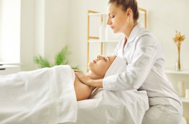 Kadın masör, güzellik salonunda ya da kozmetoloji dolabında gözleri kapalı yatan genç bir kıza omuz ve boyun rahatlatıcı masaj yapıyor. Profesyonel kozmetik uzmanı kaplıcadaki kadına masaj yapıyor..