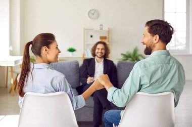 Young happy smiling couple sitting back holding hands and looking cheerful at each other during successful family therapy session with friendly male psychologist. Effective marital therapy concept.