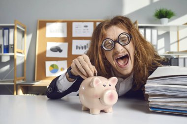 Happy crazy young man in round glasses fooling around, smiling, making funny face, having fun and putting coins in pink piggy bank. Concept of salary bonus, saving up money, investing in business
