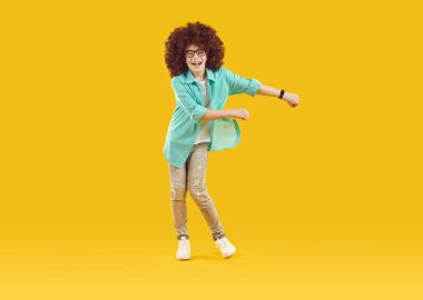 Happy cheerful playful boy in funny red curly wig, dressed in big blue shirt and gray jeans, is fooling around and dancing in good mood. Studio full-size isolated shot on yellow background.