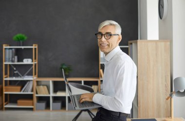 Portrait of senior businessman with laptop in hands working in modern loft office. Side view of gray-haired male manager in glasses and white shirt smiling looking at camera. Business concept.