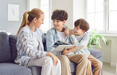 Portrait of happy family mother and her children sons sitting on couch and looking through family photo album together. Laughing young boys enjoying leisure time and good memories with their mom.