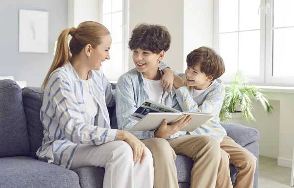 Portrait of happy family mother and her children sons sitting on couch and looking through family photo album together. Laughing young boys enjoying leisure time and good memories with their mom.