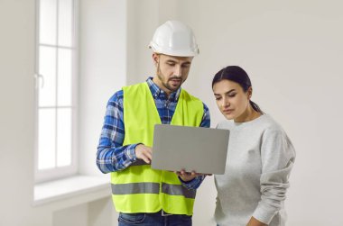 Male builder or architect meeting with lady homeowner. Young woman together with man in hard hat and uniform vest discussing new house construction and design and using modern laptop computer PC