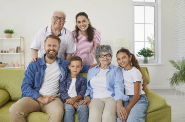 Cheerful happy family of three generations posing for family picture sitting on sofa in living room. Portrait of joyful positive grandparents with their children and grandchildren smiling at camera.