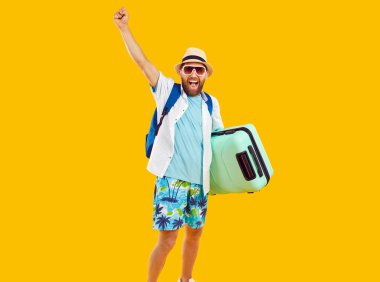 Happy man tourist in summer beach attire, isolated on orange background. With a suitcase and backpack in tow, he exudes the excitement of embarking on a well deserved vacation or holiday.