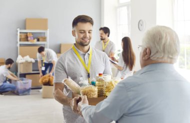 Young smiling friendly male volunteer gives a box full of grocery goods to an aged, mature, retired man in need. Charity, food donation, helping and supporting poor people concept