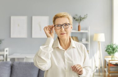 Stylish, beautiful mature woman adjusts her glasses, emphasizing her elegant demeanor. Portrait of Caucasian woman with short, styled blonde hair wearing black-framed glasses to improve her vision.