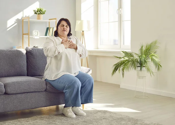 Portrait of a young plump overweight fat woman sitting on the couch in the living room at home putting a hand on her chest suffering from heart problems. Obesity problems and healthcare concept.