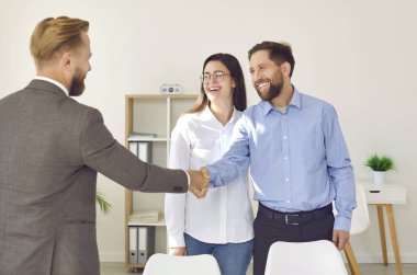 Happy couple shaking hands with real estate agent. Joyful, smiling young man and woman meeting with professional realtor, making deal, exchanging handshakes and thanking him for service