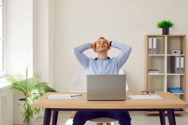 Young businessman or corporate worker completes big business project. Happy man sitting at working desk with laptop computer, holding hands behind head, feeling satisfied and smiling. Success concept