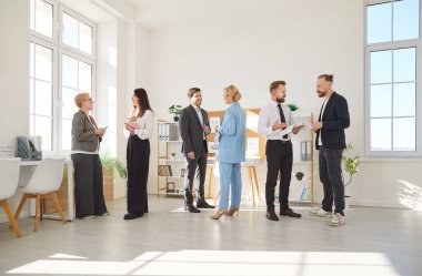 In modern office environment, several diverse groups of professionals interact during workday. People are dressed in business attire and engage in lively discussions, exchanging ideas and opinions.