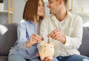 Happy married couple at home putting coins in piggy bank, spouses saving money, planning future family budget, making investment, payment for first, dreaming about holiday trip, loving together 