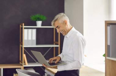 Successful businessman using laptop computer. Portrait of happy senior man standing by office desk, holding portable notebook PC, looking at screen, working on online business project and smiling
