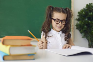 Back to school concept. A smart girl in glasses smiling writes with a pencil in a notebook while sitting at a table in the classroom. Learning education lecture task equation for children in school.