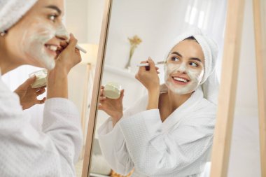 Reflection in mirror of beautiful young woman with brush applies cosmetic mask for face. Close up of woman in bathrobe and towel holding container with cosmetic product during morning beauty treatment