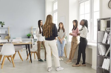 Five multiracial professional women, corporate marketing department employees engaged in briefing event, training session standing in circle in modern office, share ideas, search solution. Teamwork