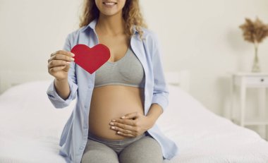 Red paper heart in hand of calm and happy pregnant woman who is in third trimester of pregnancy. Concept of expectation of pregnancy, preparation for pregnancy and childbirth. Cropped image.
