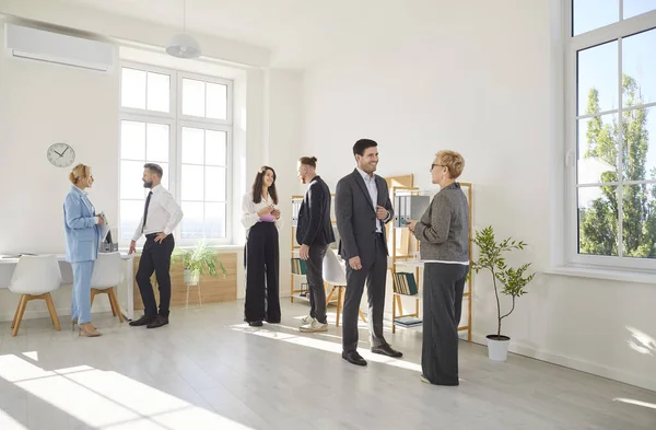 Diverse group of colleagues communicate and collaborate, creating friendly atmosphere in office. People stand in small groups discussing various topics. Business, office work and coworking concept.