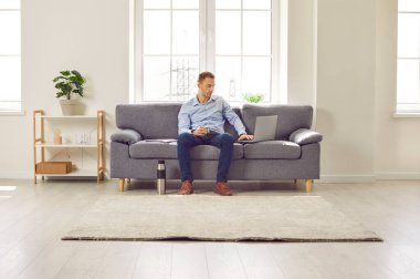 Man working from home. Busy man sitting on sofa in living room interior, using modern laptop computer, writing business emails, and drinking hot coffee from thermo bottle as he has no time for breaks