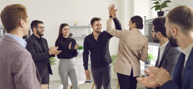 Two young men doing high five while their cheerful coworkers applauding them at meeting. Colleagues celebrating achievement, victory, successful deal in conference room in office