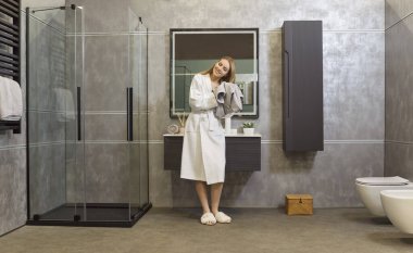 Young woman wearing white bathrobe drying hair with towel standing in bathroom interior after taking shower. Relaxed happy female person enjoying morning routine at home. Hygiene and beauty concept