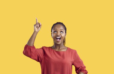 Excited joyful dark-skinned woman points her finger at copy space isolated on yellow background. Cheerful happy young casual woman in red blouse smiling looking up at content. Banner.